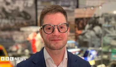 A person wearing glasses and a blue jacket stands in front of a display of racing cars, karts and motorsport memorabilia.