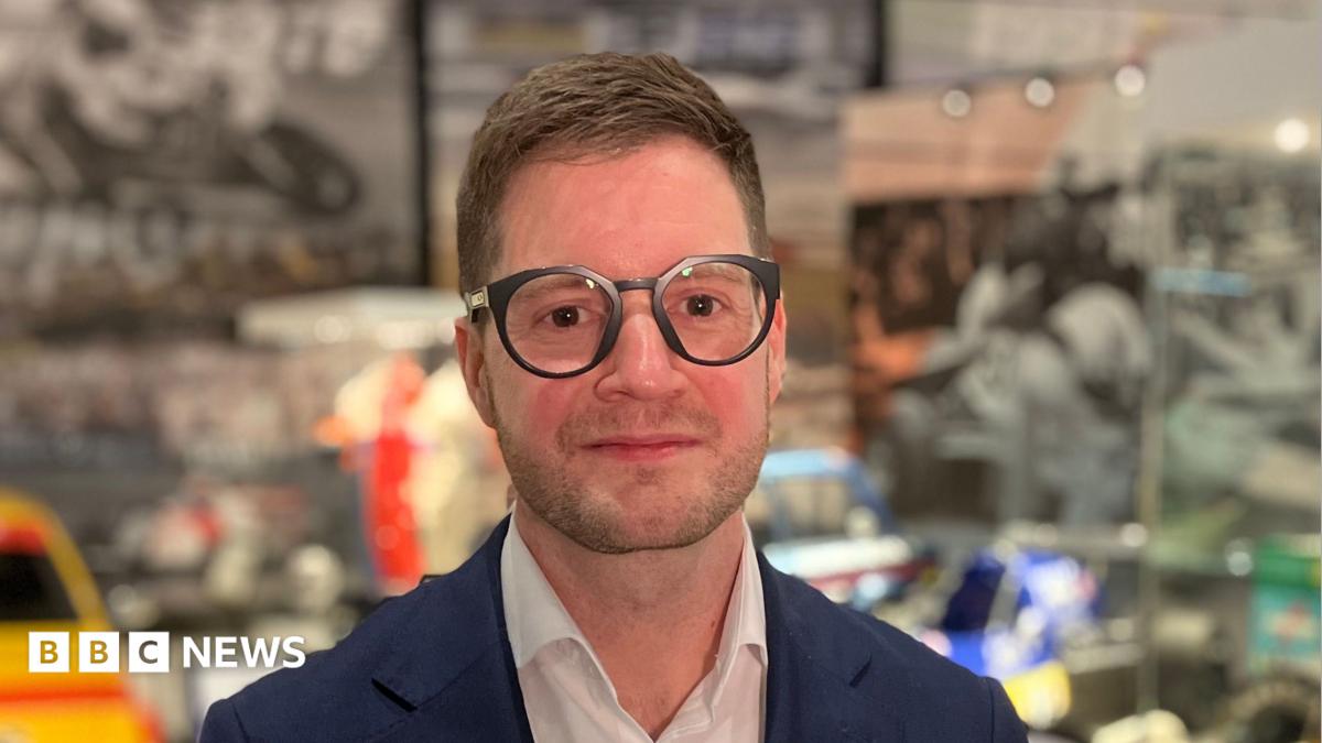 A person wearing glasses and a blue jacket stands in front of a display of racing cars, karts and motorsport memorabilia.
