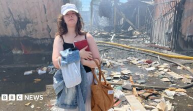 A young woman clutches a book to her chest and looks sad. Behind her is a bombed-out market structure. Destroyed books litter the ground around her