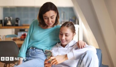A mum and daughter sit together on a blue sofa in a bright room, looking at a smartphone. The mum, who has dark brown hair and wears a turquoise top with blue jeans, has her arm around her daughter's shoulder. The daughter has fair hair tied back and wears a grey hoodie and holds the phone. A kitchen with white cupboards, blue wall tiles, wooden worktops, chairs, appliances and a fruit bowl is blurred in the background.