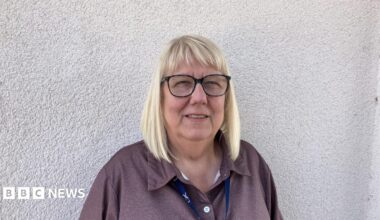 Linda has shoulder length blonde hair and is waring glasses. She is standing by a white wall which is behind her and is wearing a lanyard around her neck.
