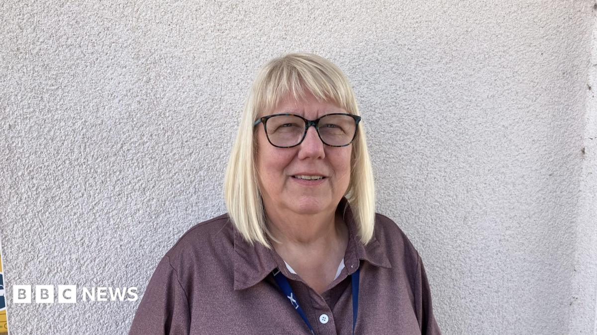Linda has shoulder length blonde hair and is waring glasses. She is standing by a white wall which is behind her and is wearing a lanyard around her neck.