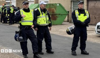 Three police officers are pictured standing in a road. They are wearing yellow vests and all have caps on