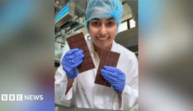 A woman holding two bars of chocolate while smiling into the camera. She is wearing a white gown with purple gloves and a blue hair net.