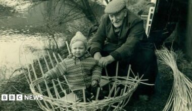 Black and white photo, an elderly man in a flat cap making a round boat out of straw with a baby sat inside along the banks of the river.