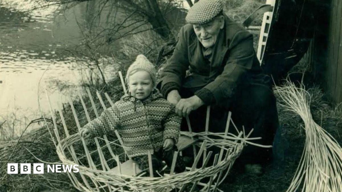 Black and white photo, an elderly man in a flat cap making a round boat out of straw with a baby sat inside along the banks of the river.