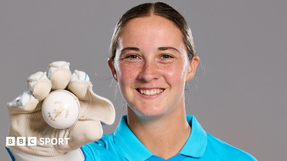 A smiling Alice Capsey holds up a ball while wearing wicketkeeper gloves