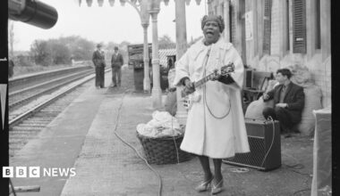 Sister Rosetta Tharpe stands on th epaltform in a large coat playing an electric guitar  on the station platform.