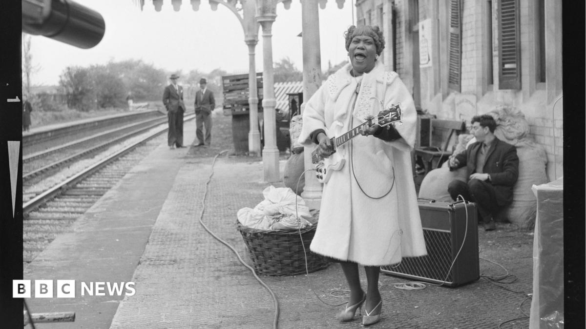 Sister Rosetta Tharpe stands on th epaltform in a large coat playing an electric guitar  on the station platform.