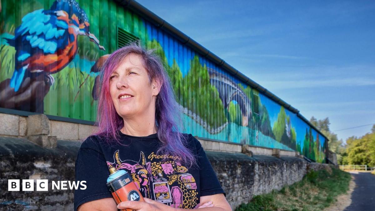 A woman stands in front of a colourful mural and holds a spray can. She has purple hair and glances off to the side
