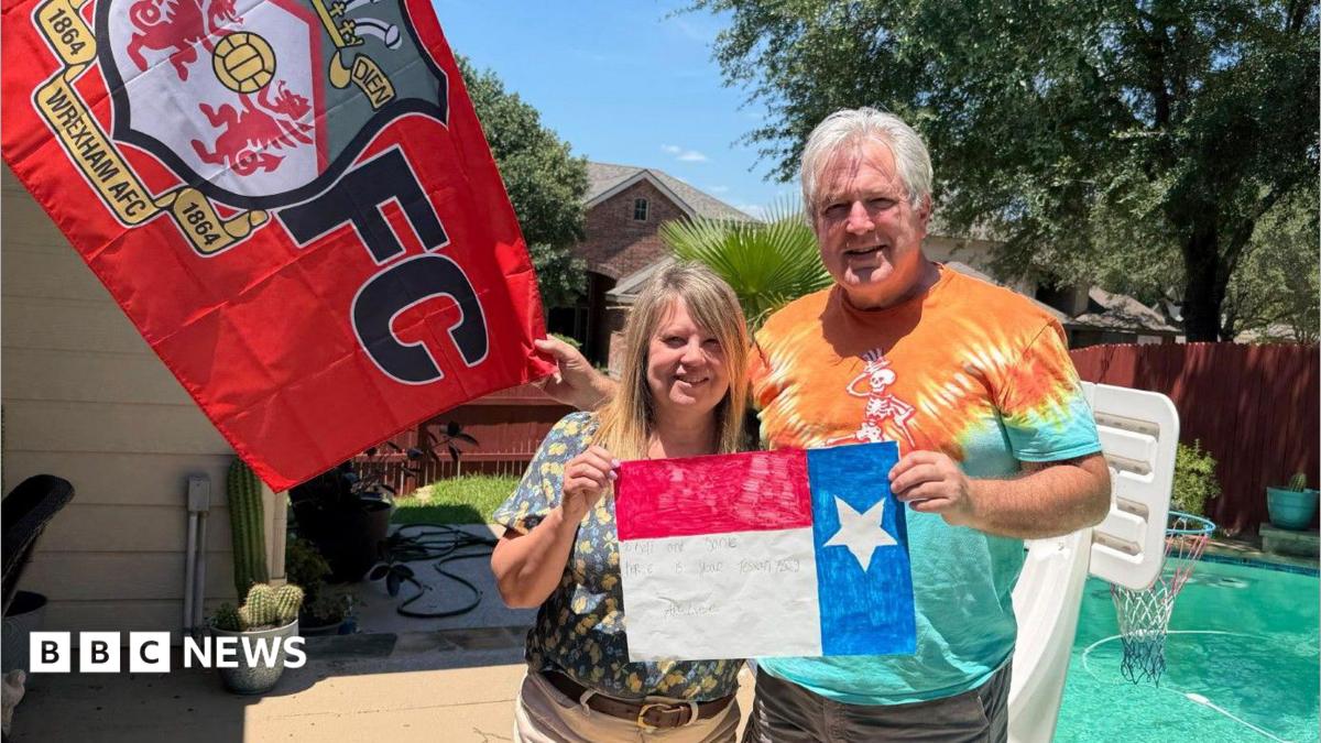 A man and a woman in their early 60s, wearing shorts and t-shirts, stand in the sunshine in front of a small back garden swimming pool. They are holding a painting of the Texas flag between them and there is a  large Wrexham AFC flag behind them to the left. The woman has blonde shoulder-length hair and the man has short white hair. They are both smiling. Trees, plants, a fence and a neighbouring house are visible in the background.
