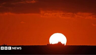 The Sun sets over an ocean. The sky is dark red and the silhouette of a ship sailing across the ocean in front of the Sun.