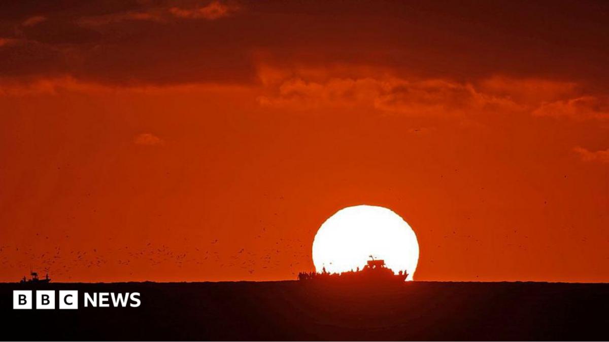 The Sun sets over an ocean. The sky is dark red and the silhouette of a ship sailing across the ocean in front of the Sun.