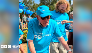 David Treasure in a blue Parkinson's UK t shirt with a Parkinson's UK cap and sunglasses. He has his arms outstretched towards his walker which is out of shot. There are people to the side of him and behind and a balloon arch with Parkinson's UK balloons.