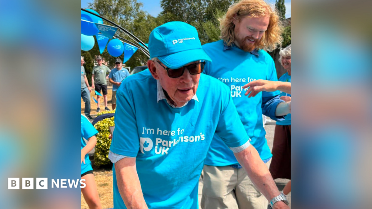 David Treasure in a blue Parkinson's UK t shirt with a Parkinson's UK cap and sunglasses. He has his arms outstretched towards his walker which is out of shot. There are people to the side of him and behind and a balloon arch with Parkinson's UK balloons.