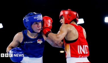 Ruby White and Sakshi Chaudhary wearing protective headgear exchange punches in a boxing ring. White wearing blue representing England is struck by an Chaudhary in red representing India under bright arena lights.