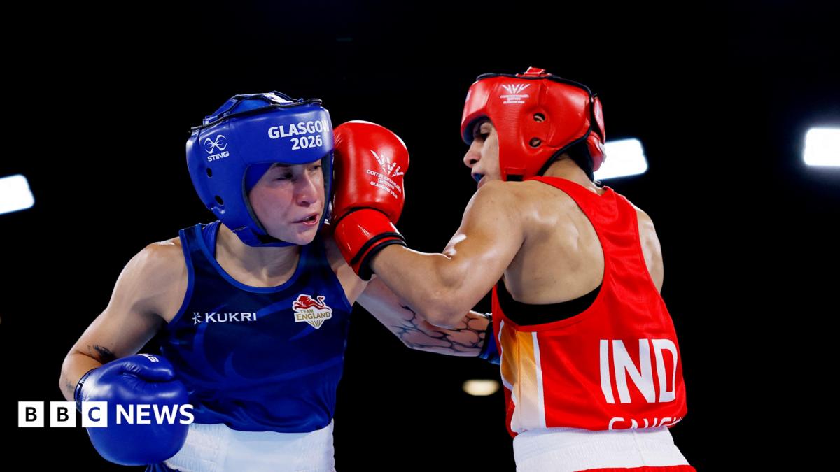 Ruby White and Sakshi Chaudhary wearing protective headgear exchange punches in a boxing ring. White wearing blue representing England is struck by an Chaudhary in red representing India under bright arena lights.