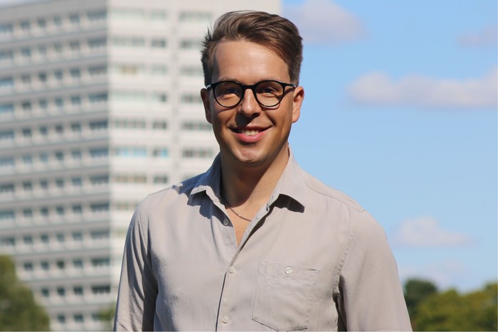 ben-wadey Ben Wadey stands in front of a tower block. He is wearing a grey shirt and glasses and is smiling