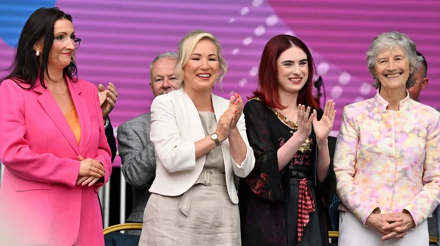 Four women are standing together. The two on the outside have their hands clasped. The two in the middle are clapping. All are smiling. Other people and a large screen and chairs are in the background.