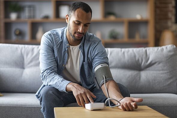 Black Man Sitting On Couch, Checking Blood Pressure With Upper Arm Monitor Black Man Sitting On Couch, Checking Blood Pressure With Upper Arm Monitor