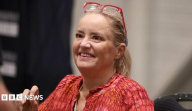 Lucy Davis, a fair-haired woman in a red top and with red-framed glasses worn on top of her head, is seen sitting down and smiling at the For The Love of Fantasy & Sci-Fi convention at Excel London on Saturday 22 August 2026