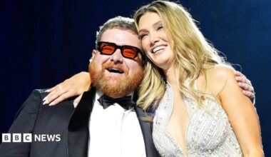 A smiling Cammy Barnes, who is wearing a dinner suit, is hugged by Delta Goodrem, who is wearing a silver sparkly dress. Cammmy has ginger hair and beard and black framed glasses with red lenses. Delta has long blonde hair.