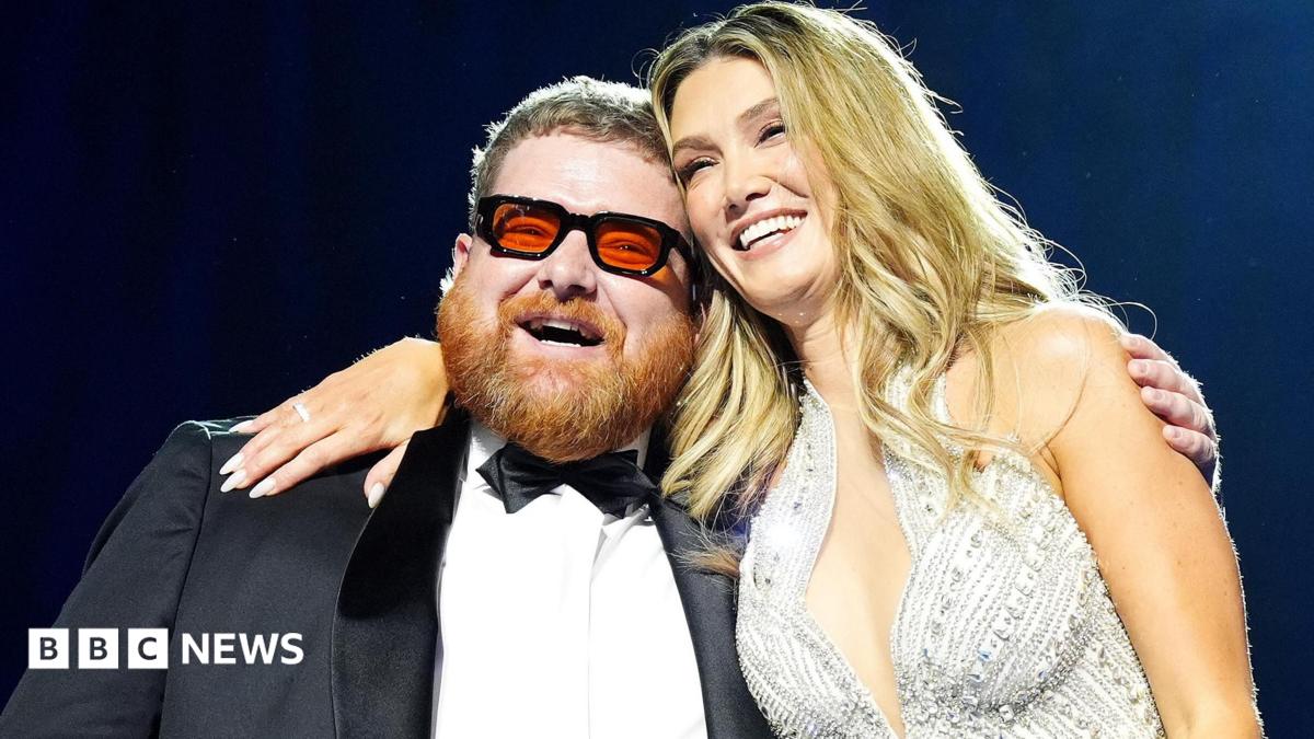 A smiling Cammy Barnes, who is wearing a dinner suit, is hugged by Delta Goodrem, who is wearing a silver sparkly dress. Cammmy has ginger hair and beard and black framed glasses with red lenses. Delta has long blonde hair.