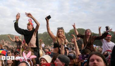Crowds of festival-goers wave their hands in the air at Leeds Festival. There a few people in the crowd raised up by sitting on the shoulders of other people.