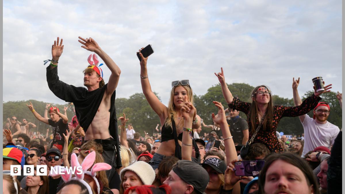 Crowds of festival-goers wave their hands in the air at Leeds Festival. There a few people in the crowd raised up by sitting on the shoulders of other people.