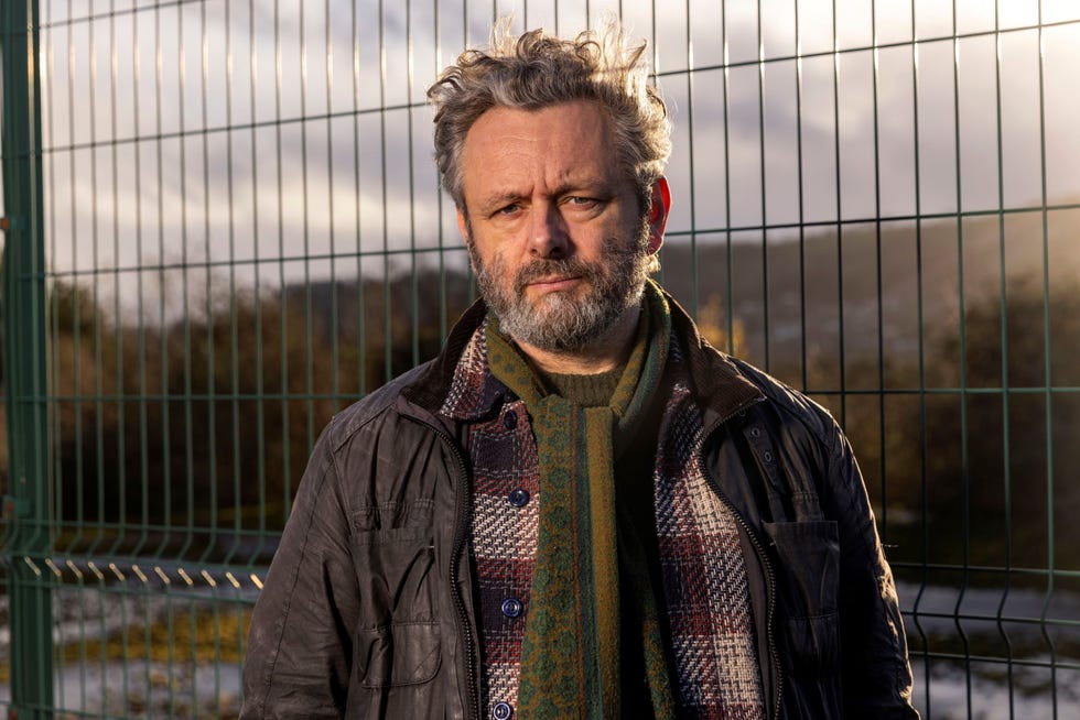 buried michael sheen man with a beard standing in front of a metal fence, wearing a scarf and jacket.