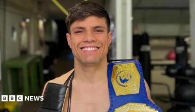 A man with dark hair smiling with a boxing belt over his shoulder.