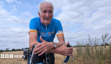 An older man with white hair is on a bicycle in a field of crops. He is wearing a blue cycling t-shirt that reads "hope house children's hospices"