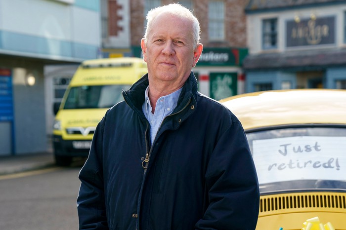 Charlie Fairhead on Casualty poses for the camera. He has a smile on his face and is dressed in a blue shirt and blue jacket. He is stood in front of an ambulance and a yellow car which has a sign on it, reading "just retired"