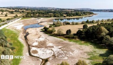 An aerial image of Chew Valley Lake shows the water level is exceptionally low, with much of the dry lake bed exposed. The lake is surrounded by brown fields.