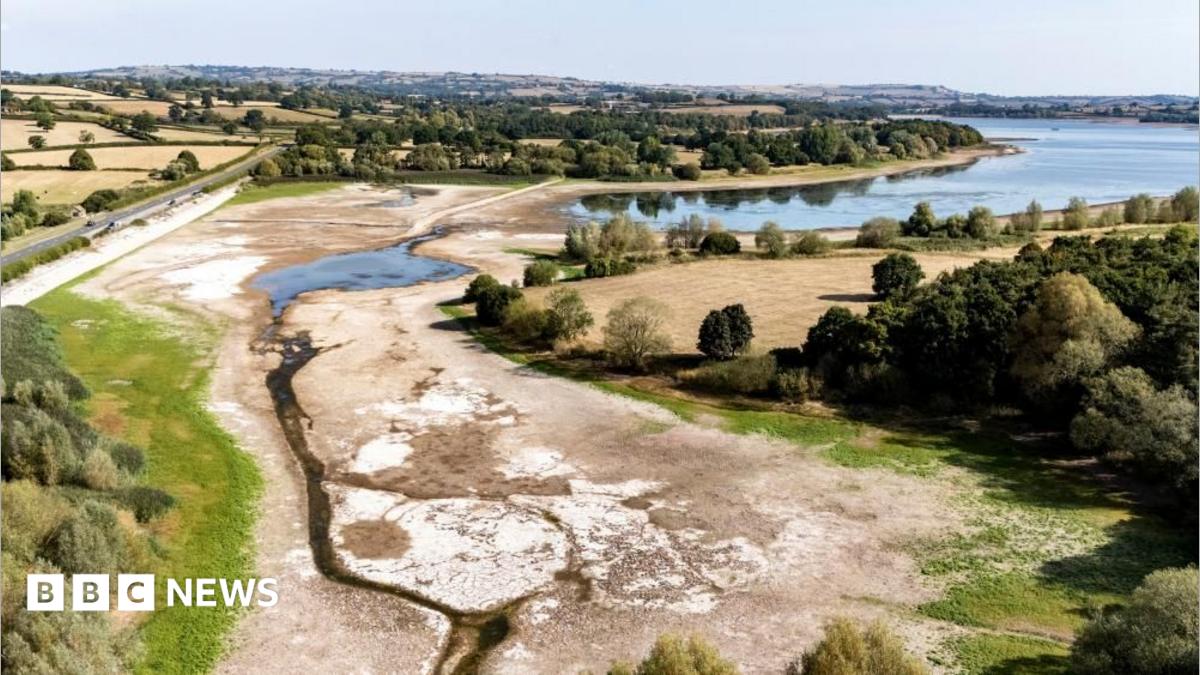 An aerial image of Chew Valley Lake shows the water level is exceptionally low, with much of the dry lake bed exposed. The lake is surrounded by brown fields.