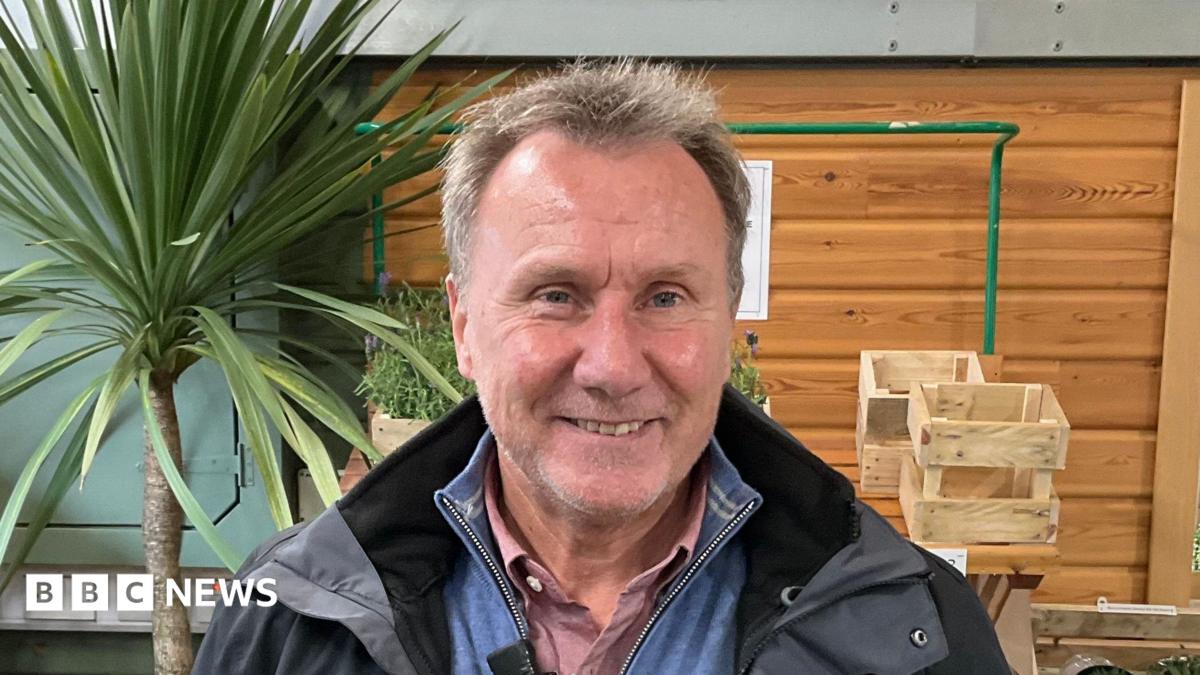 Peter Gould, a man with short grey hair and stubble. He is wearing a salmon coloured shirt underneath a light blue quarter zip jumper. He is also wearing an unzipped, black raincoat which features the Jersey prison service crest. Behind him, to the right of the image, are a series of wooden boxes on a shelf, and a mirror on the wall. To the left of the image is a palm tree, and a set of wooden shelves on which there are transparent plastic bags of kindling wood.