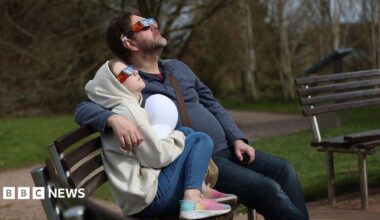 A man and a child watching a partial solar eclipse at Keele university on 29 March, 2025. They are wearing special glasses and are sat on a bench and there is part of what looks like an empty bench next to them. There is grass in the background.