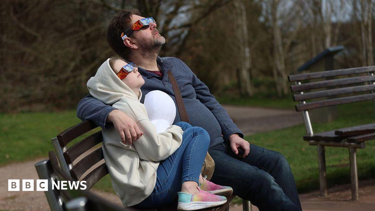 A man and a child watching a partial solar eclipse at Keele university on 29 March, 2025. They are wearing special glasses and are sat on a bench and there is part of what looks like an empty bench next to them. There is grass in the background.