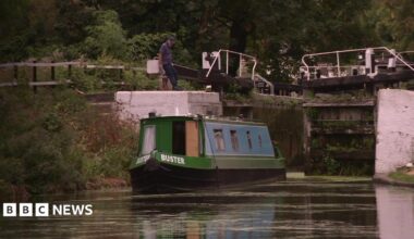 A narrowboat on a canal approaches a lock, while a person stands beside the lock gates. Trees and vegetation line the waterway in the background.