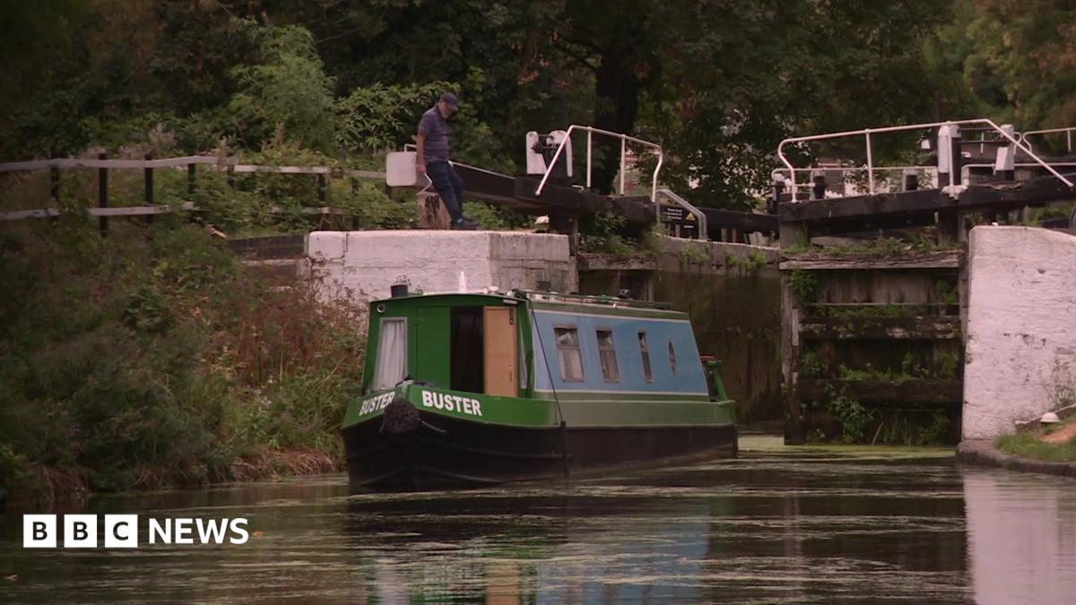 A narrowboat on a canal approaches a lock, while a person stands beside the lock gates. Trees and vegetation line the waterway in the background.
