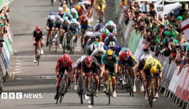 A peloton of cyclists racing on a road, surrounded by crowds of people cheering from behind barriers.