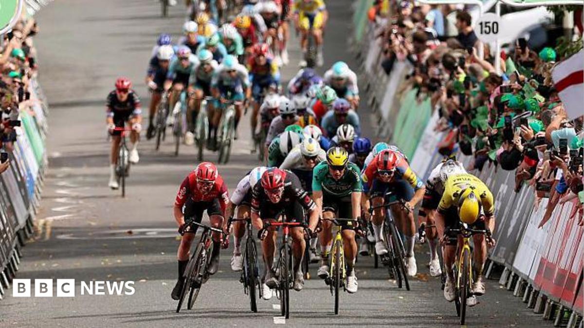 A peloton of cyclists racing on a road, surrounded by crowds of people cheering from behind barriers.