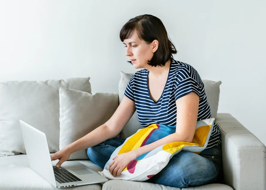 An image of a woman sitting on a grey couch with a pillow on her lap, looking at a laptop set beside her while having a virtual meeting with her nutritionist.