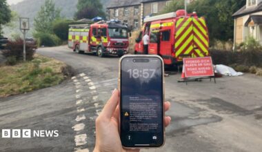 A phone screen with an emergency alert on is seen on a phone, with fire engines behind