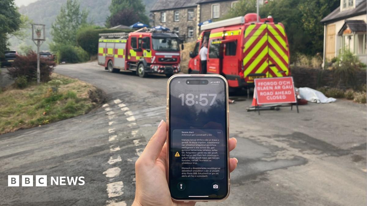 A phone screen with an emergency alert on is seen on a phone, with fire engines behind