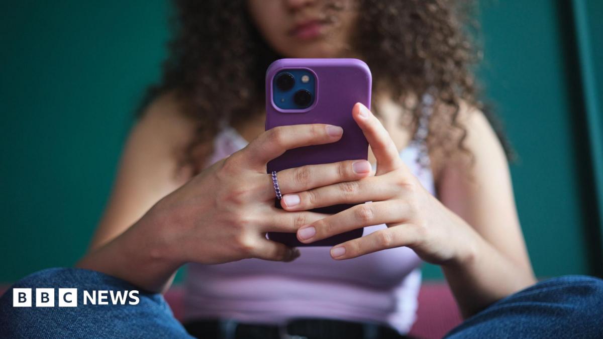 A young woman, whose face cannot be seen, sits cross-legged while holding and using her smartphone. She is wearing blue jeans and a lilac vest top.
