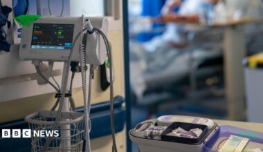 Medical equipment in a hospital. On the left of the image is a machine which measures vital observations, while on the right - on a trolley - are small bottles. In the background, out of focus, is a hospital bed through a door.