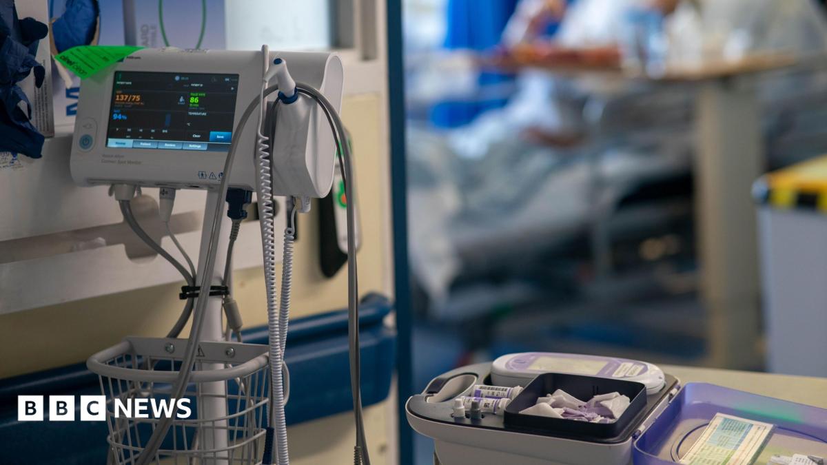 Medical equipment in a hospital. On the left of the image is a machine which measures vital observations, while on the right - on a trolley - are small bottles. In the background, out of focus, is a hospital bed through a door.