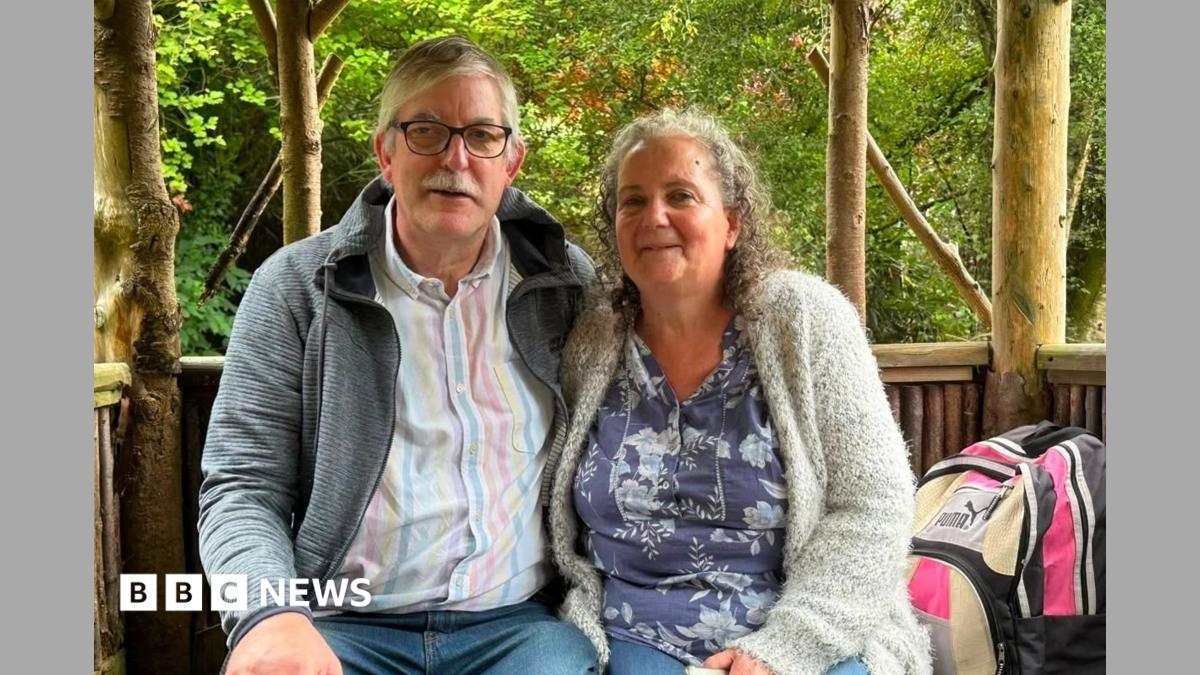 A couple in their 60s stand in front of a waterfall which has rocks and greenery around it. She is wearing a white shirt and gold chain necklace, her hair is shoulder length and clipped back from her face, and is grey and curly and she has the strap of a brown bag across her shoulder.   He has white hair and a moustache and is wearing a white shirt with a palm tree pattern.
