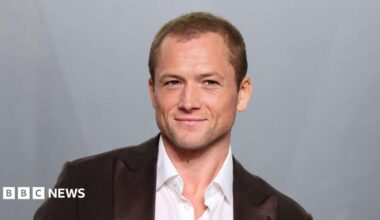 Egerton, a man in his late 30s wearing a white shirt and a brown suit jacket. There's a grey background behind him.